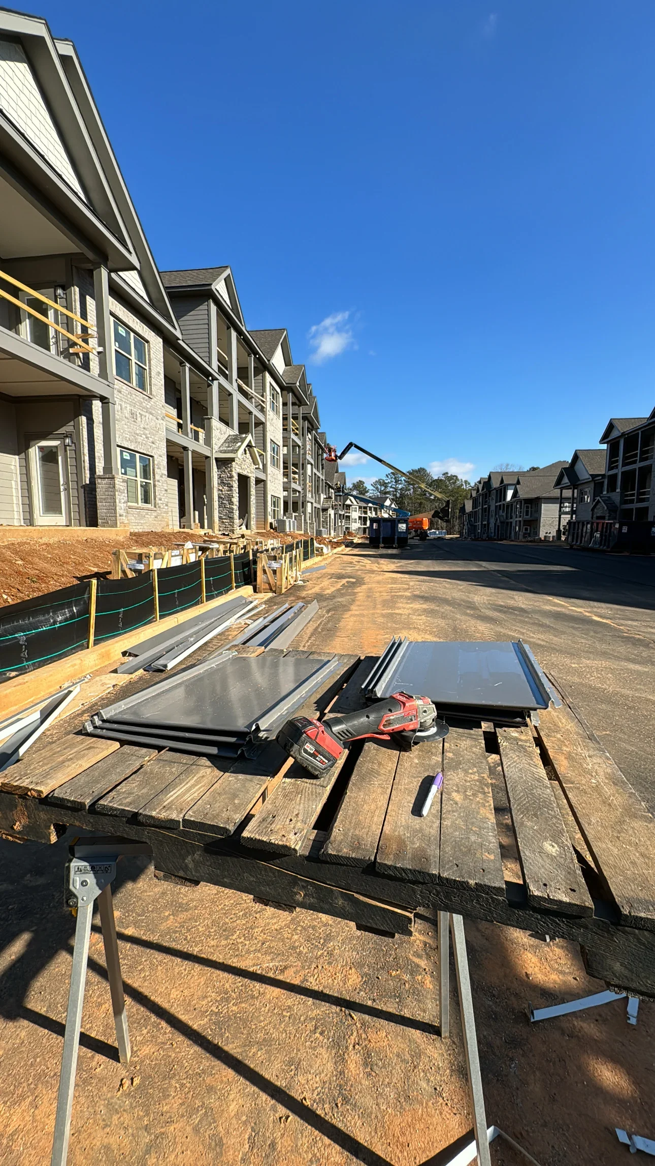 Black shingles and metal roofing materials staged on wooden pallets at residential construction site with multiple homes under development