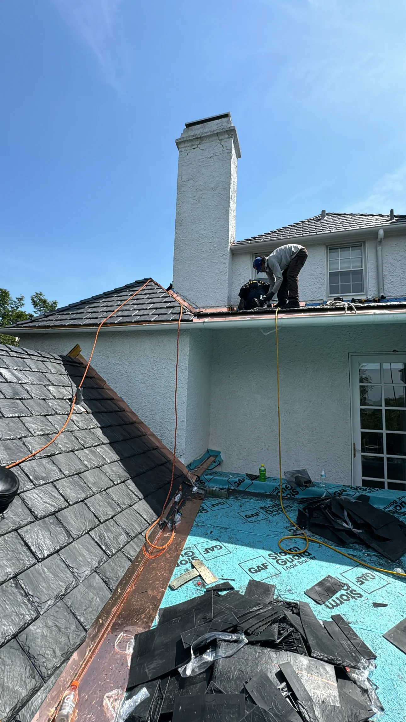 Roofer installing metal flashing around tall white chimney with slate and clay tile roof sections
